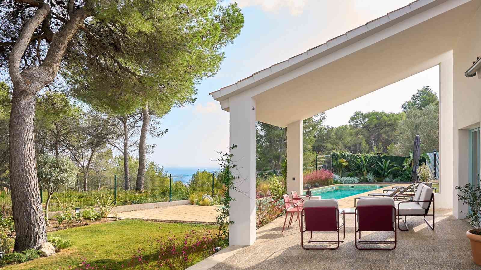 A white verandah with chairs next to a pool and greenery on a sunny day.
