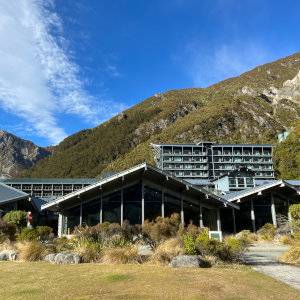 A blue-grey hotel building nestled within mountains and bushes on a sunny day.