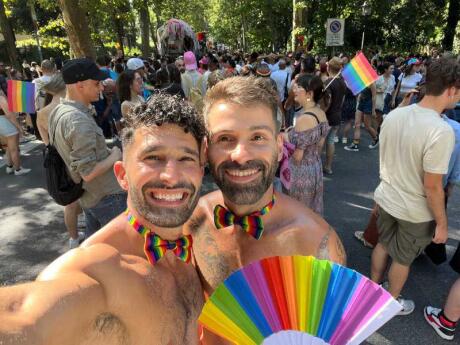 Stefan and Seby posing in rainbow bow ties and a rainbow fan at a park with more people and rainbow flags behind them.